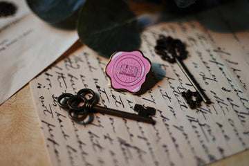 No0neDesigns Bookish Wax Seal enamel pin resting against a background of letters that were hand written and antique keys.