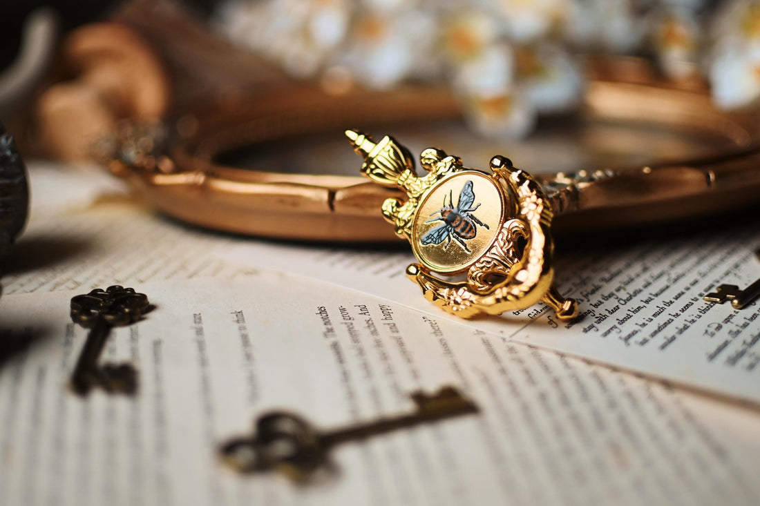 The image features the bridgerton inspired bee knocker pin by No-one designs with the moveable handle. The pin is propped against a blurred background ontop of book pages.