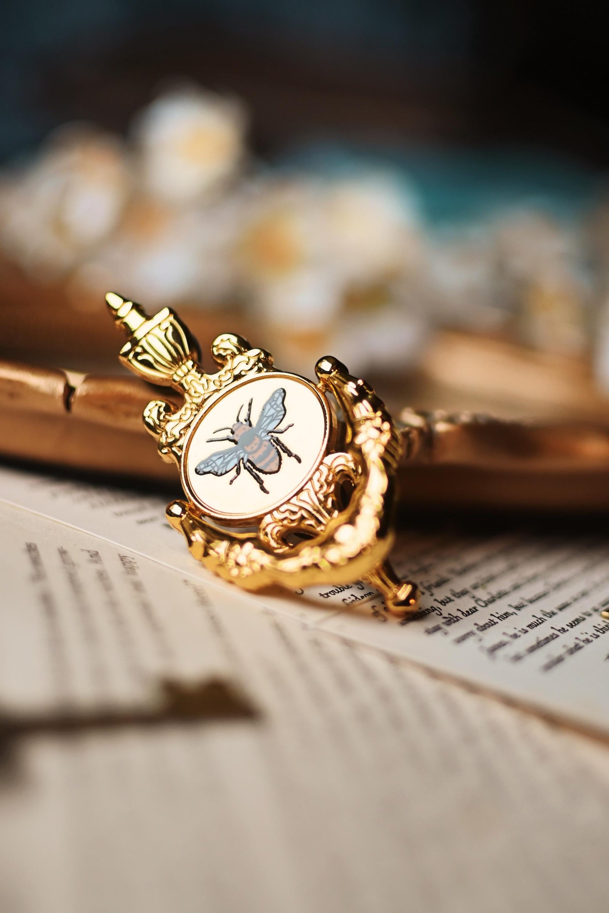 The image features the bridgerton inspired bee knocker pin by No-one designs with the moveable handle. The pin is propped against a blurred background ontop of book pages.