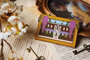 An enamel pin featuring an illustration of Bridgerton House with purple flowers and a yellow frame, placed on a surface with a vintage aesthetic.