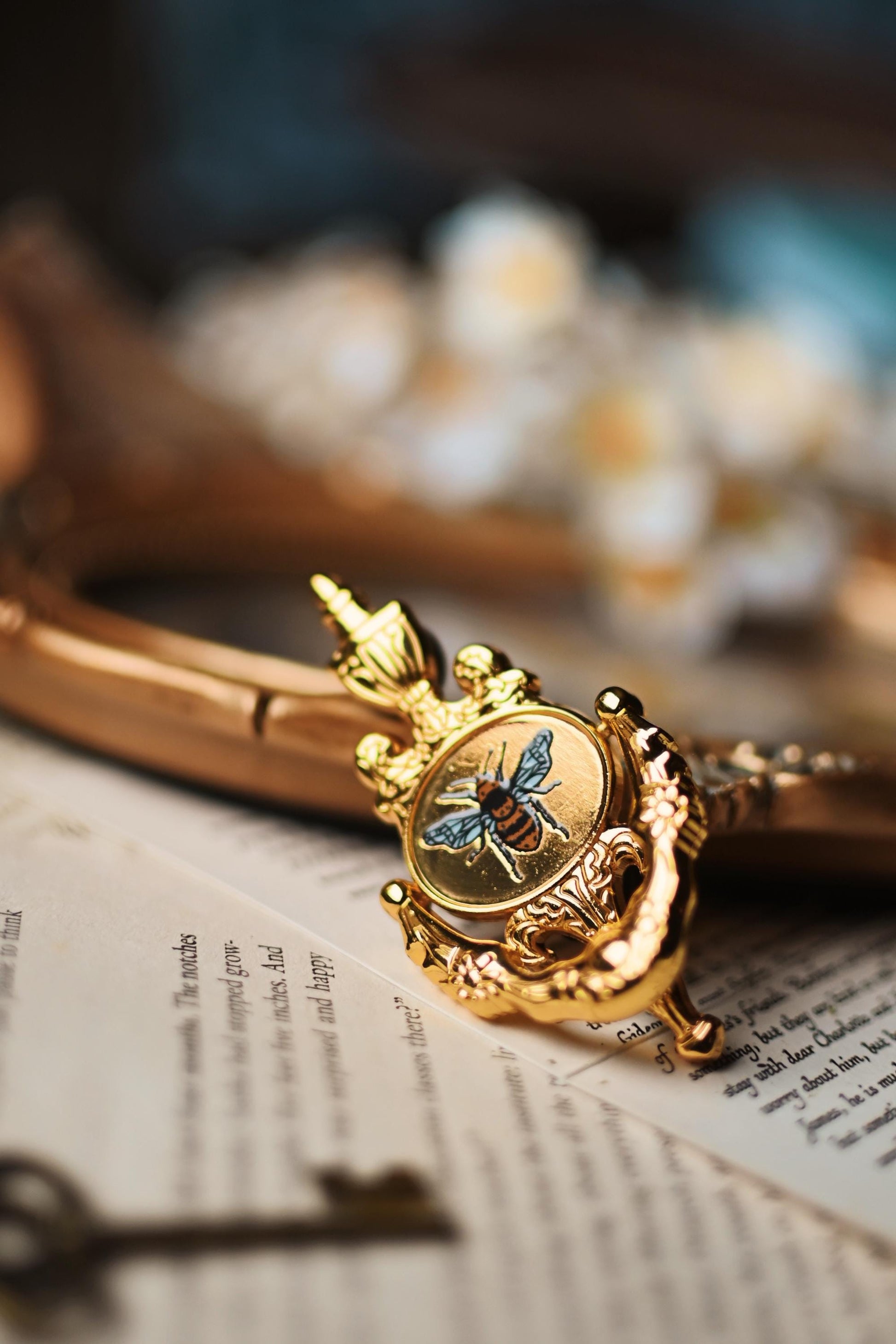 The No0neDesigns Bridgerton inspired moveable Bee Knocker enamel pin resting against a tray with a blurred background and on top of book pages. 