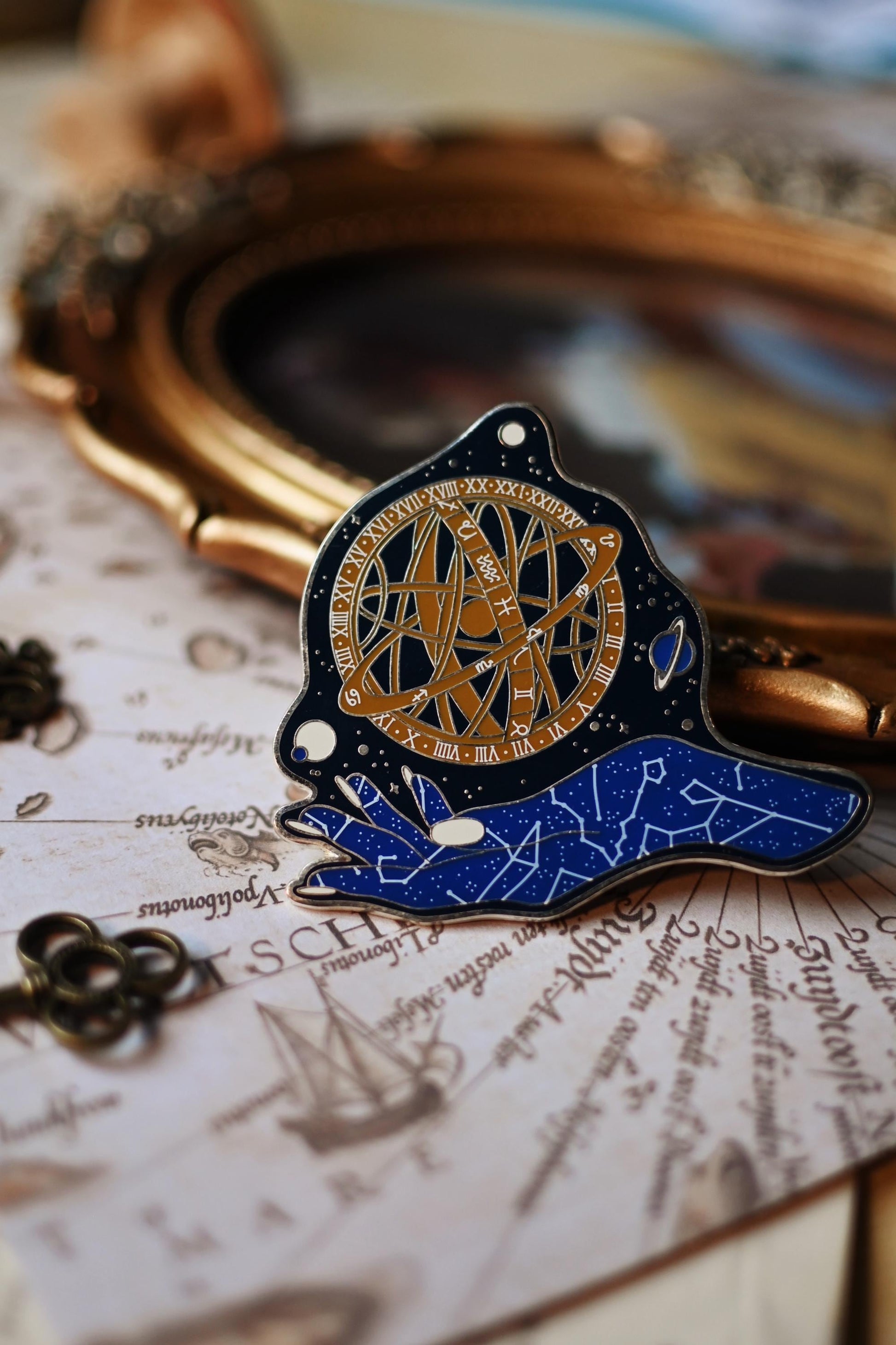 An enamel pin depicting a celestial goddess design with gold, blue, and black colors on a silver metal backing, displayed on a book with written pages.