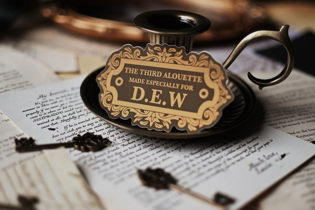A sticker with vintage design, gold and black in color, with the text 'THE THIRD ALOUETTE especially for D.E.W inspired by Rebecca Ross' Devine Rivals.