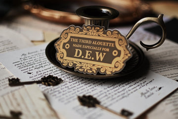 A sticker with vintage design, gold and black in color, with the text 'THE THIRD ALOUETTE especially for D.E.W inspired by Rebecca Ross' Devine Rivals.