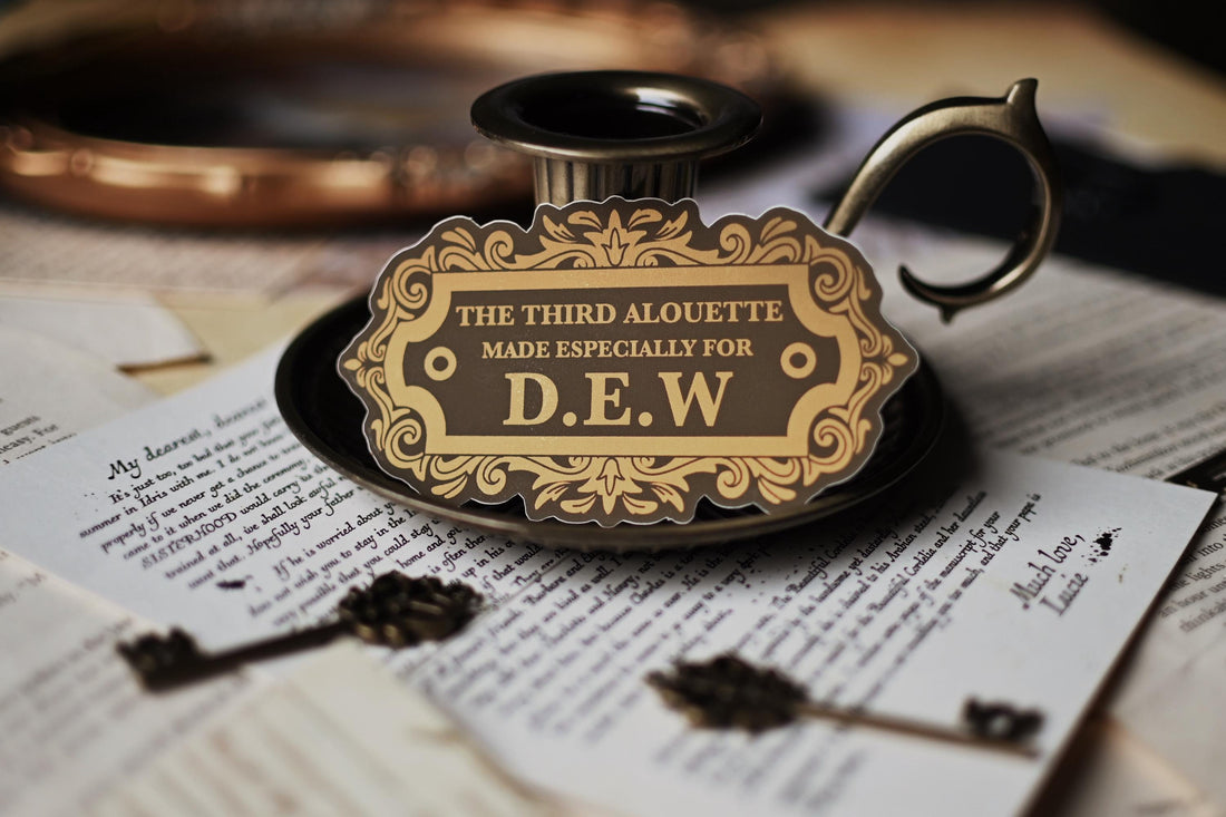 A sticker with vintage design, gold and black in color, with the text 'THE THIRD ALOUETTE especially for D.E.W inspired by Rebecca Ross' Devine Rivals.