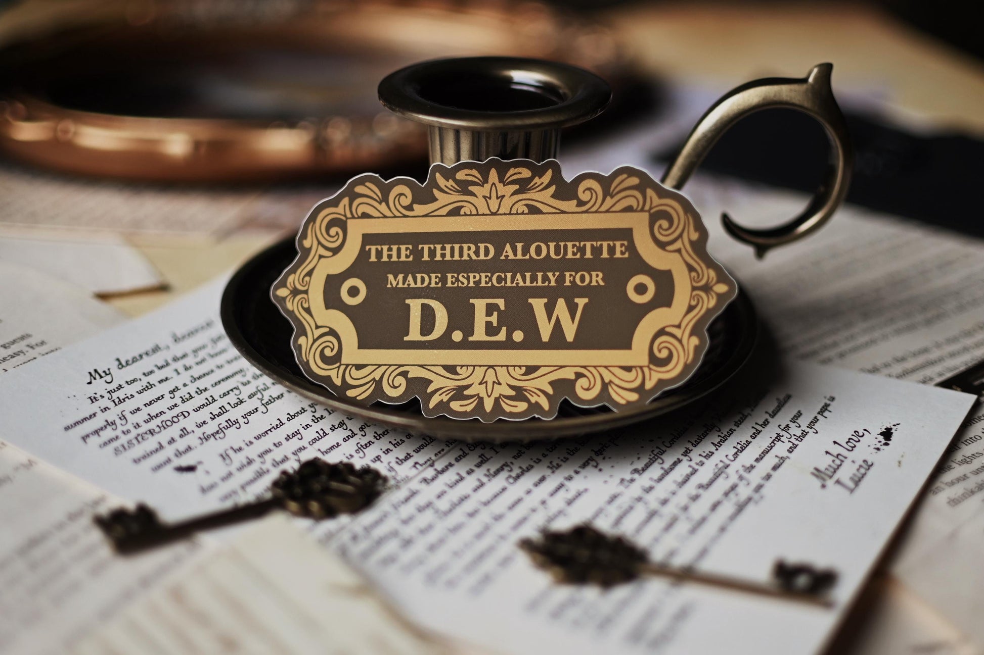 A sticker with vintage design, gold and black in color, with the text 'THE THIRD ALOUETTE especially for D.E.W inspired by Rebecca Ross' Devine Rivals.