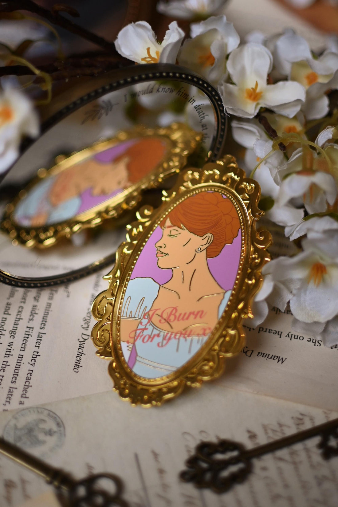 Daphne Bridgerton illustration printed onto an enamel pin in the style of a mirror with the quote I Burn For You written on top. The mirror pin rests against an actual mirror while resting on top of bookpages. 