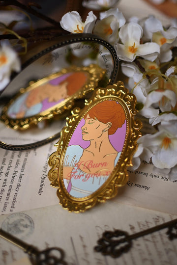 Daphne Bridgerton illustration printed onto an enamel pin in the style of a mirror with the quote I Burn For You written on top. The mirror pin rests against an actual mirror while resting on top of bookpages. 