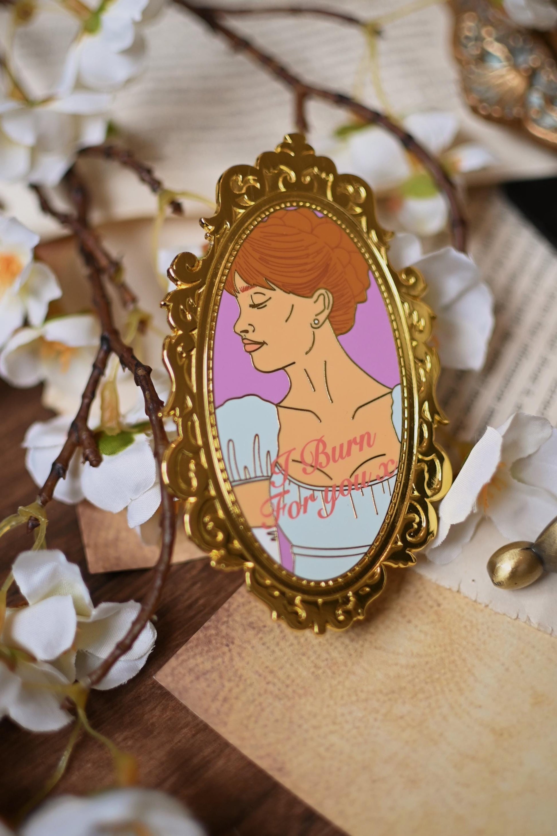 No0neDesigns Daphne Bridgerton Mirror enamel pin inspired by the Bridgerton series. The image showcases the mirror pin that features Daphne Bridgeton resting against a background of orchids and bookpages. 
