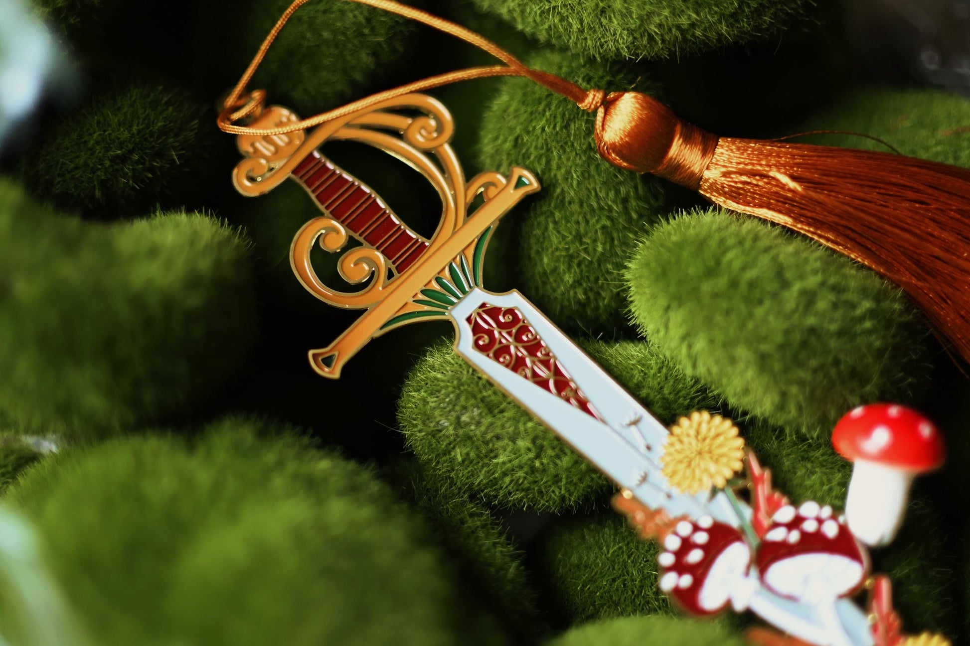 A close up shot of The Fae Queen bookmark featuring a bookmark made of soft enamel in the shape of the fae queen sword on top of a bed of fake moss. 