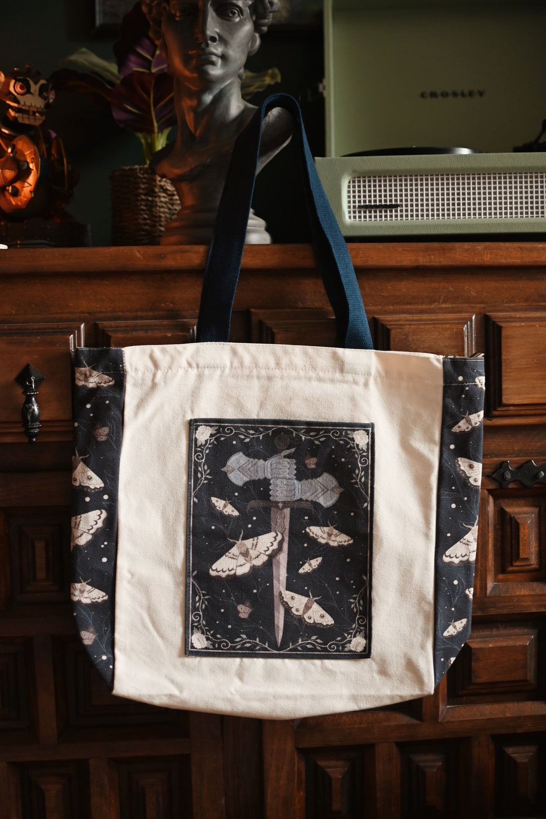 The Knight and the Moth inspired tote bag with decorative design hanging on a wooden cabinet.
