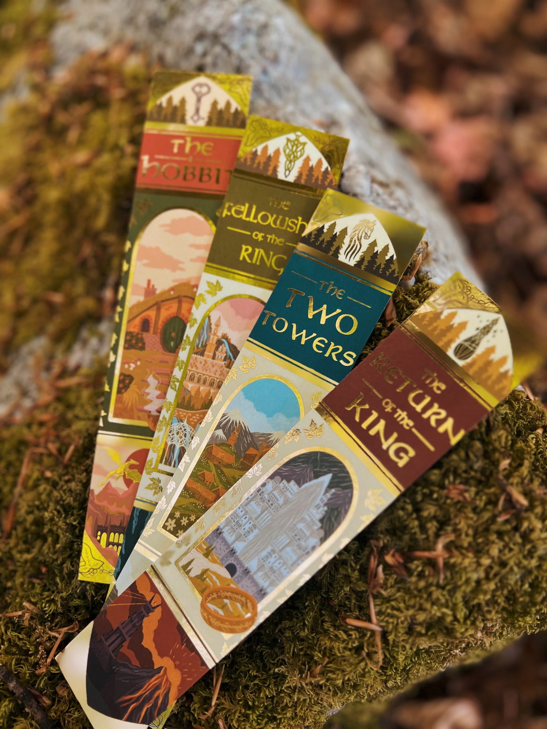 Taken outside and featuring No0neDesigns Lord of the Rings and the Hobbit inspired foil bookmark collection fanned out on top of a tree branch, resting on a bed of moss.