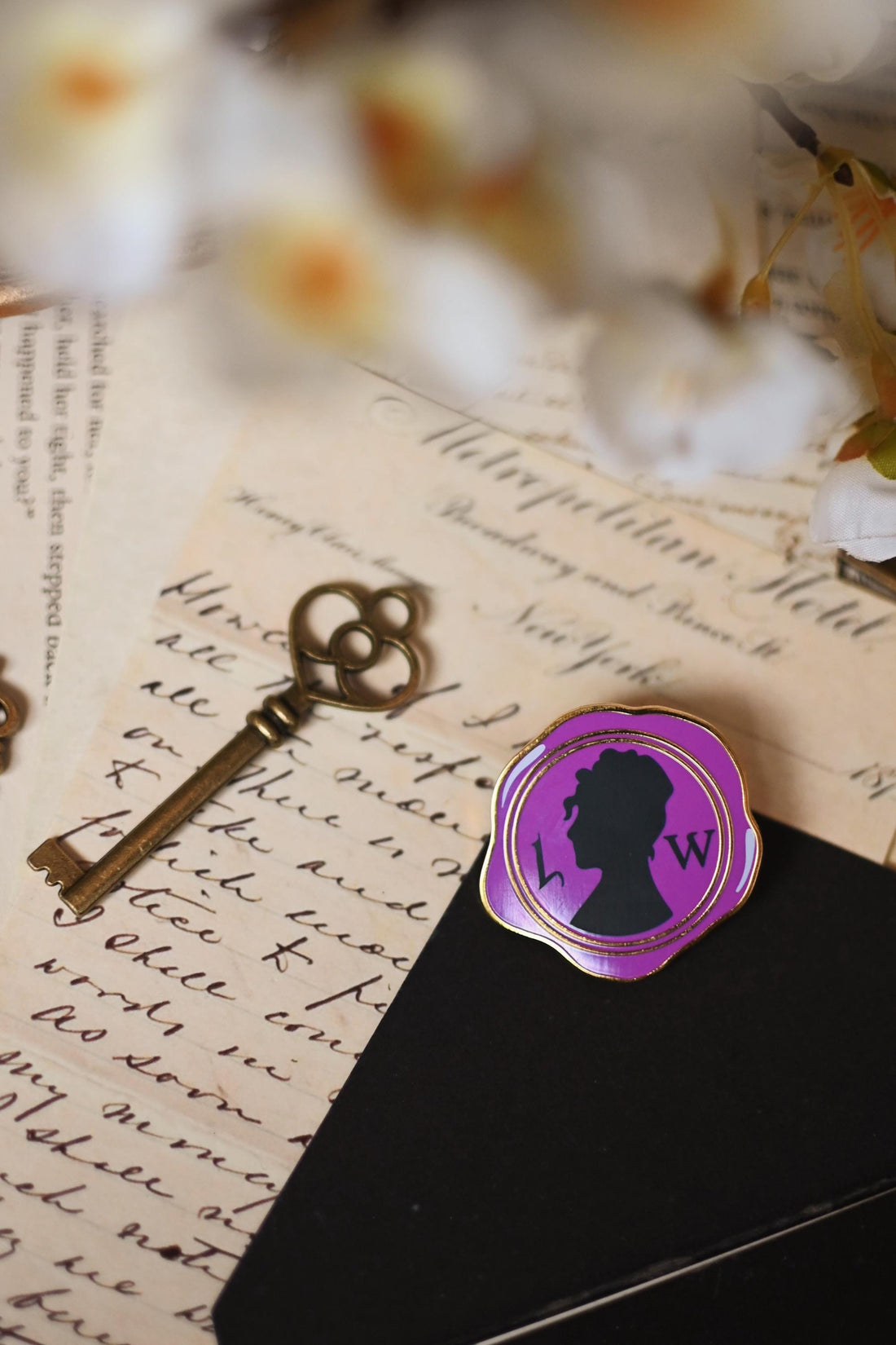 An enamel pin with a purple silhouette of a woman's profile, inspired by Lady Whistledown, placed on a black surface with a vintage background including a key and floral elements.