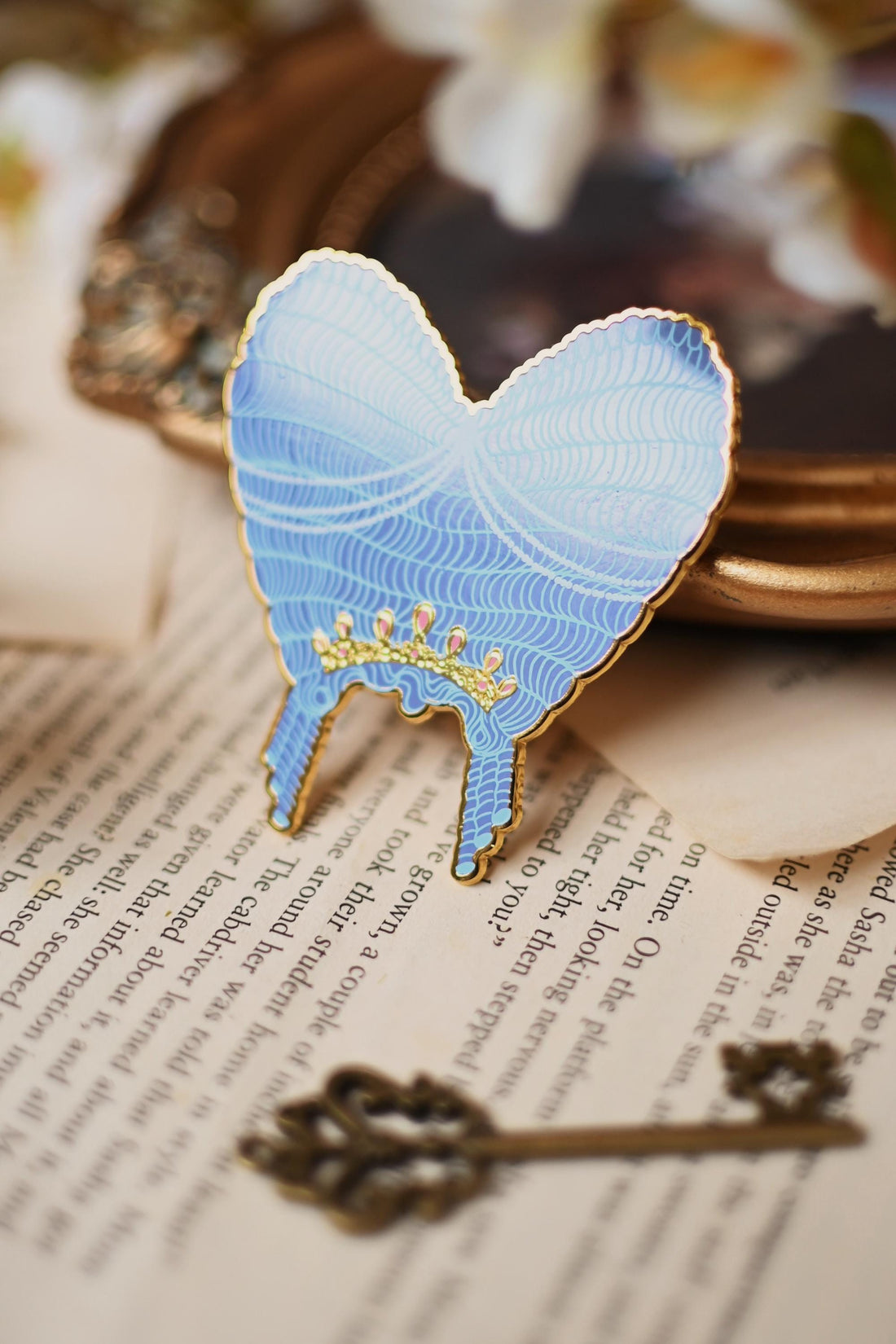 No0neDesigns Bridgerton Collection featuring the Queen Charlotte Wig enamel pin. The pin is help upright against a tip of a mirror to show the silk pint detailing of the wig and crown detailing too.