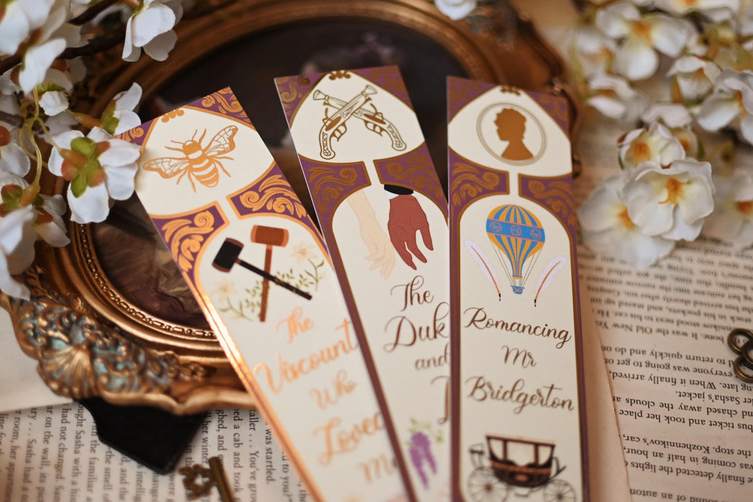 No One Designs Bridgerton-inspired bookmark collection with the three bookmarks fanned out against bookpages and a mirror that raised the bookmarks into the foreground. Each bookmark is inspired by one of Julia Quinn's books and features a gold-foil detailing. Bookmarks inspired by The Duke & I, The Viscount Who Loved Me and Romancing Mr Bridgerton. 