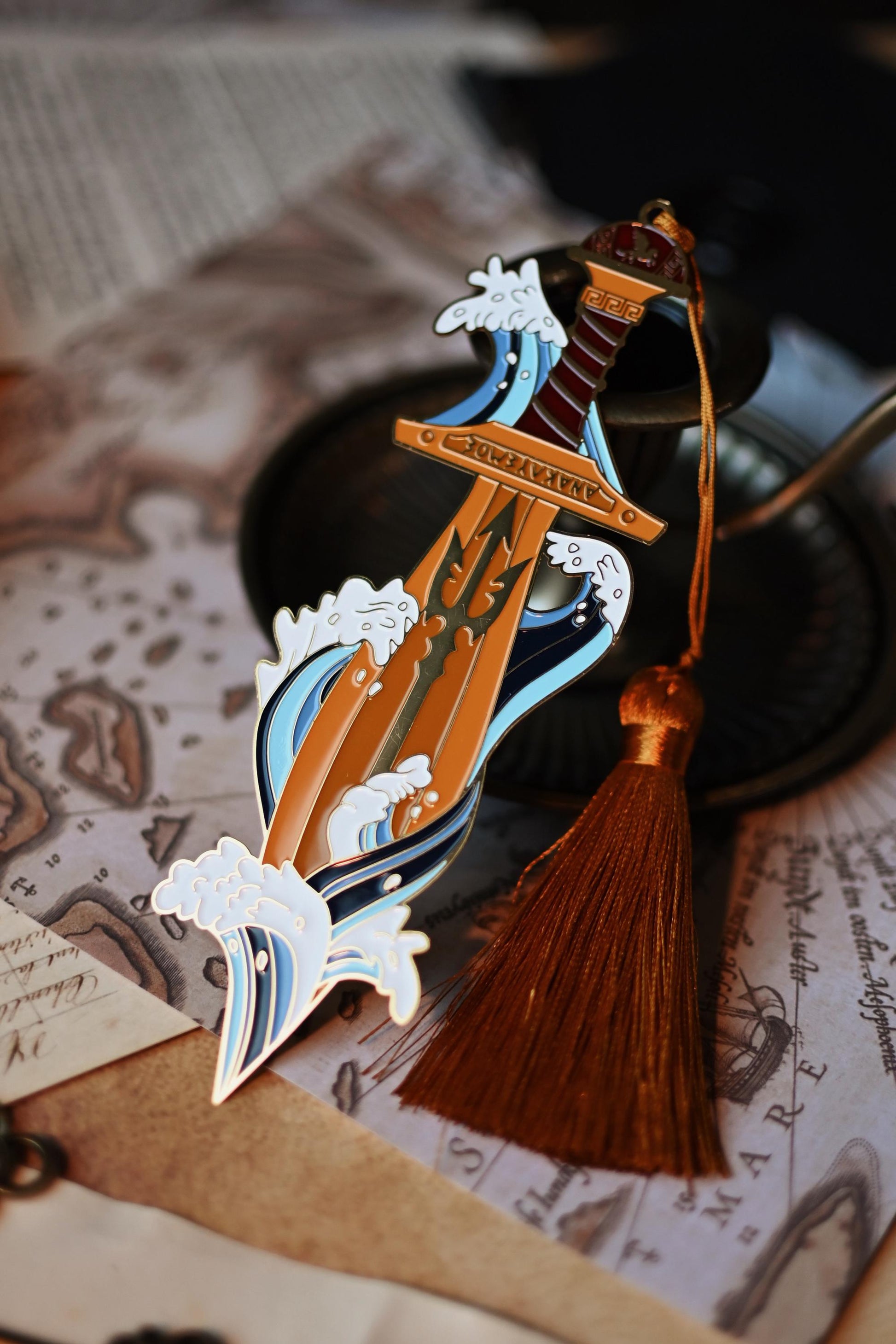 Percy Jackson inspired enamel pin featuring Percy's sword riptipe surounded by ocean waves. The bookmark is propped up against a candleholder and shines against the light to showcase a trident design in the sword. 