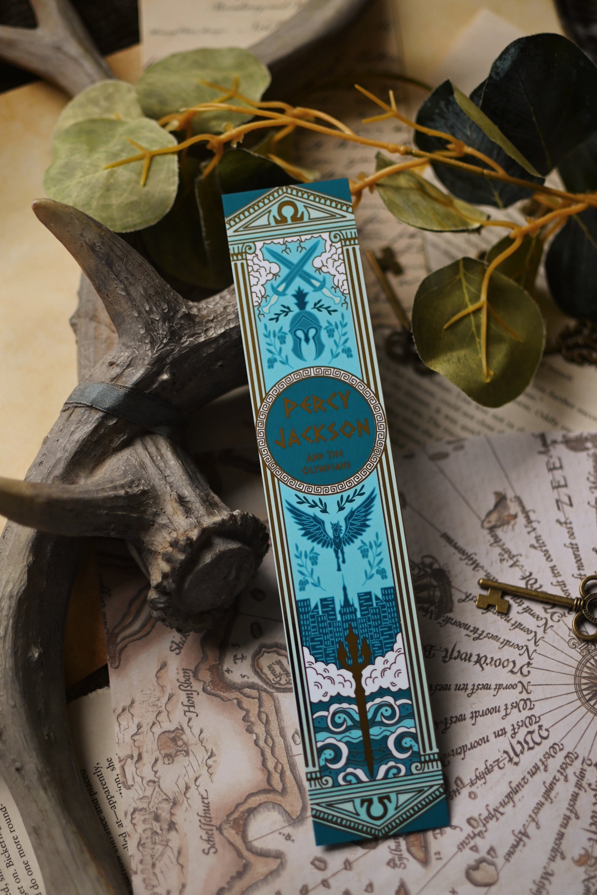 The Percy Jackson Book Spine inspired decorative bookmark with blue and gold design resting on a map with antlers and keys in the background