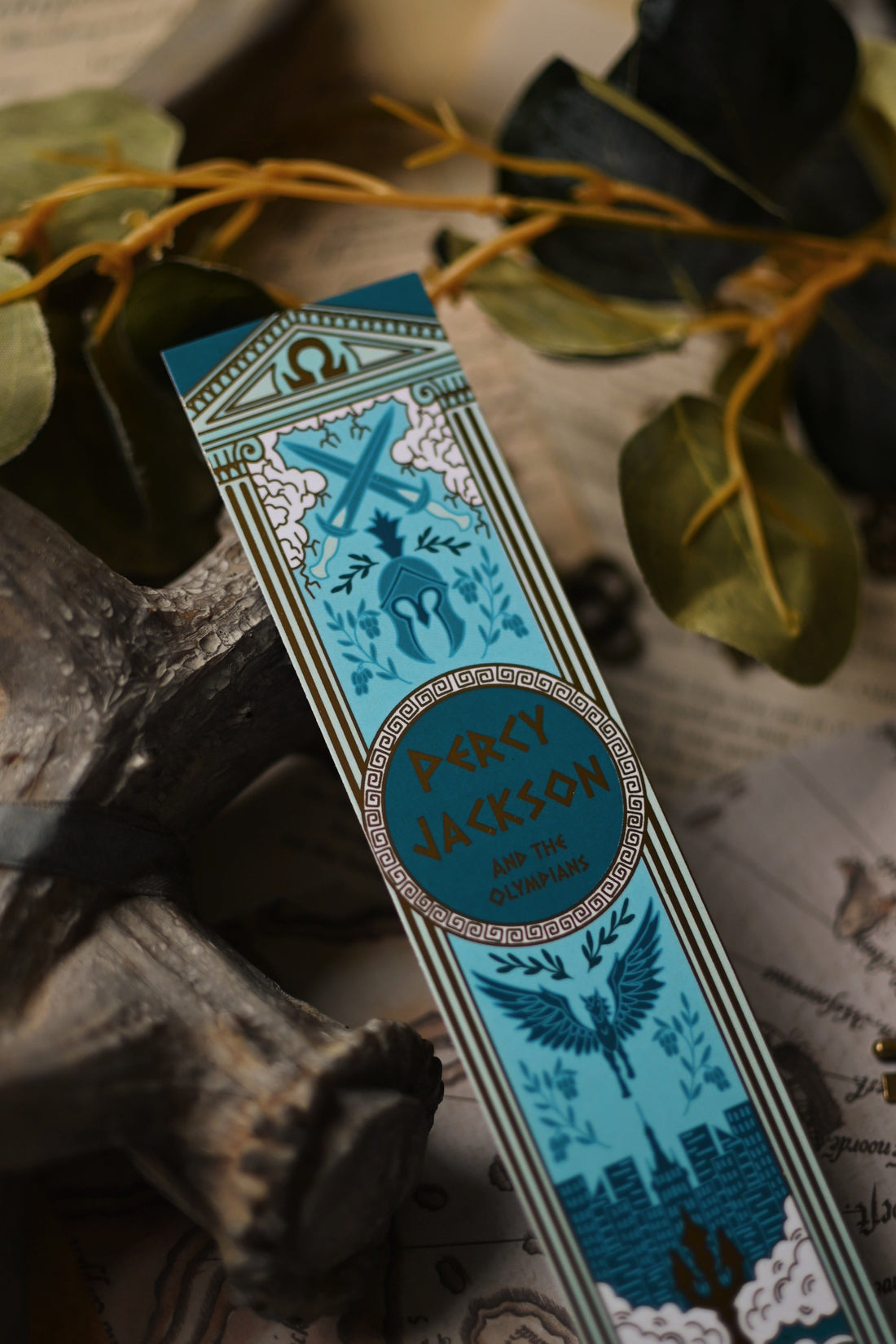 The Percy Jackson Book Spine inspired decorative bookmark with blue and gold design resting on a map with antlers and keys in the background