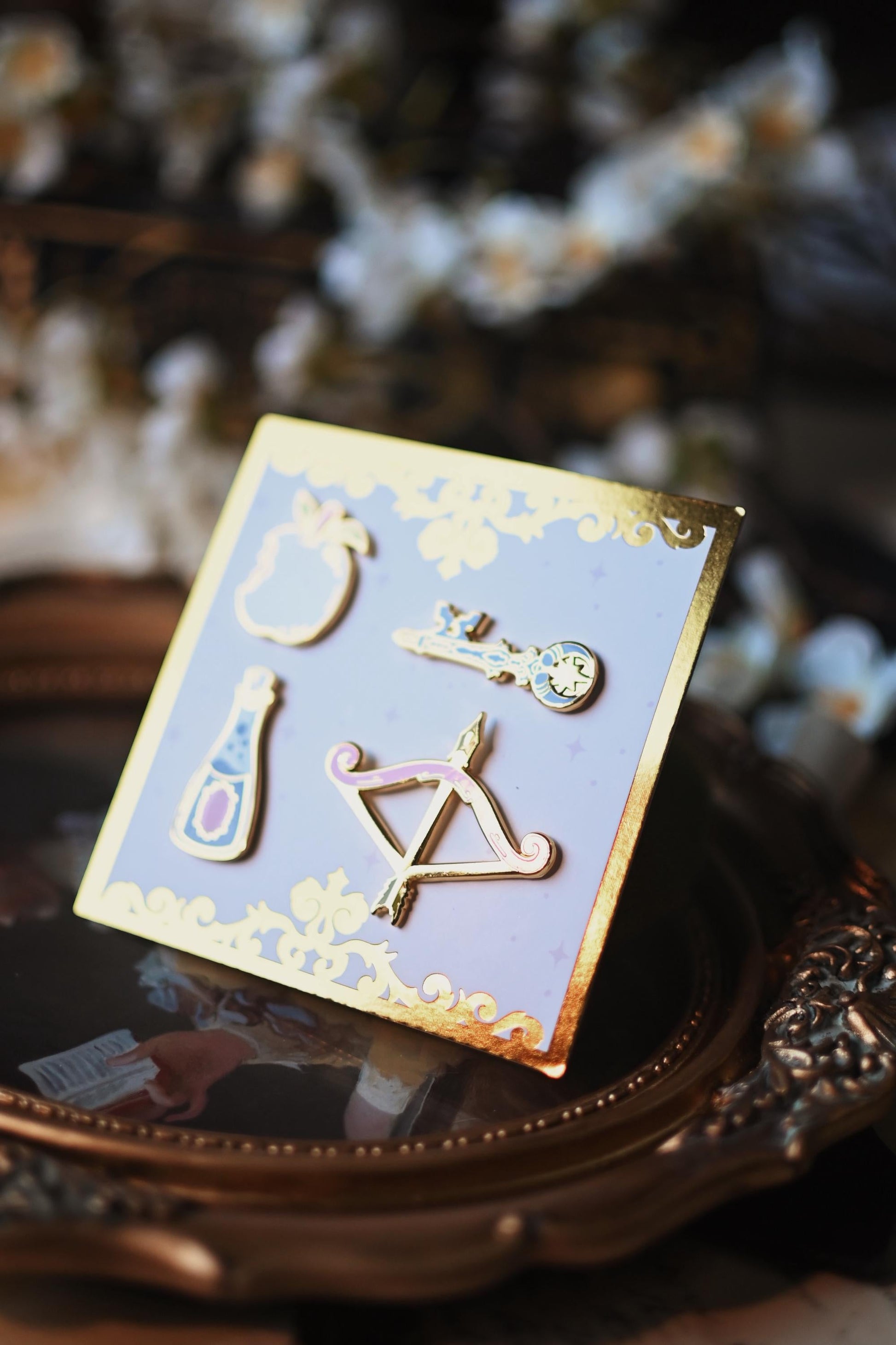 A set of four pins with intricate designs displayed on a card, positioned on a vintage-style surface with floral motifs. The pins are directed towards the light to showcase the foiling and gold of the pin. 