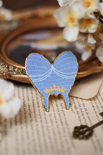No0neDesigns Bridgerton Collection featuring the Queen Charlotte Wig enamel pin. The pin is help upright against a tip of a mirror to show the silk pint detailing of the wig and crown detailing too.