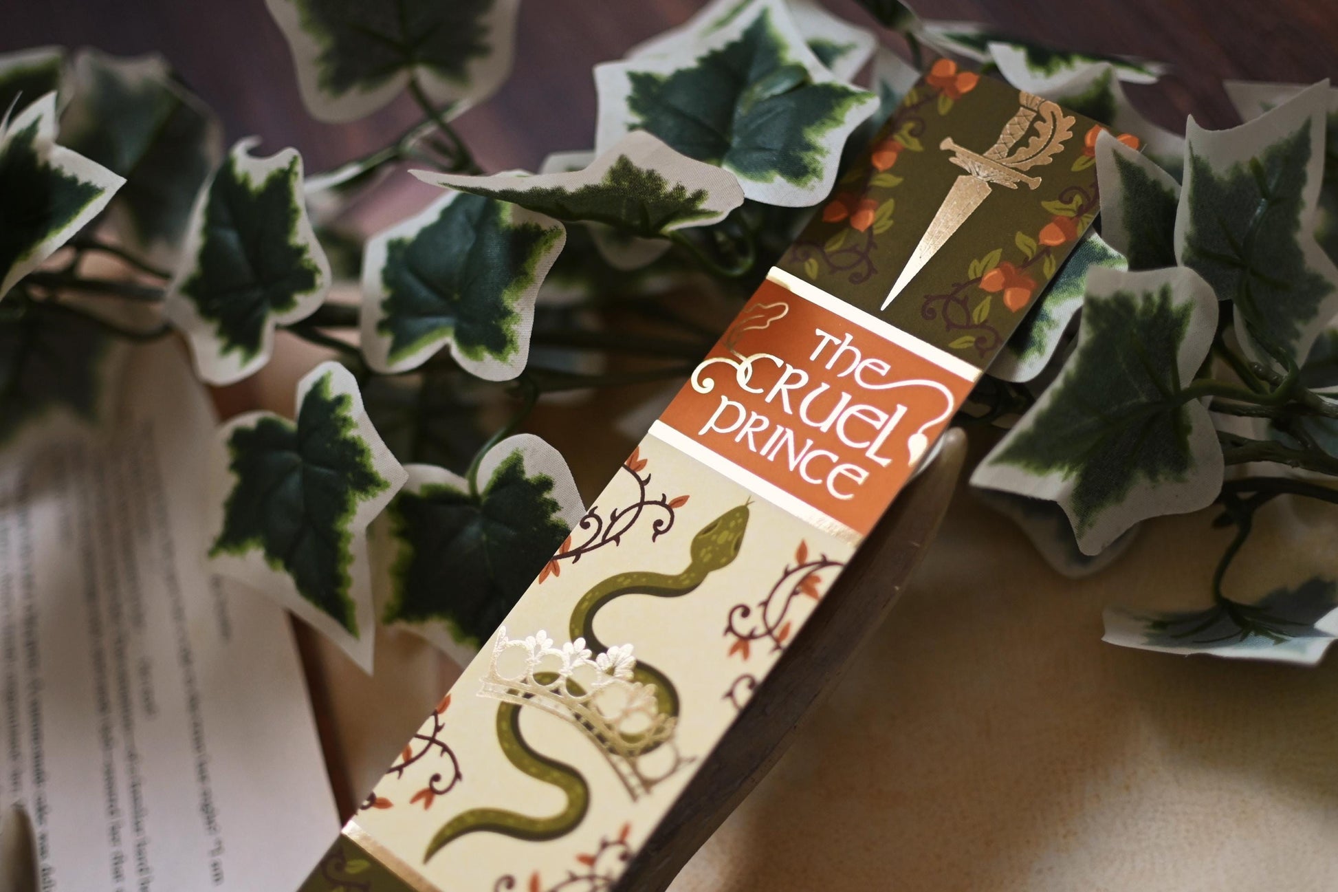 No One Designs book spine bookmark featuring a design inspired by The Cruel Prince by Holly Black. The bookmark is tilted up towards the camera to showcase the gold detailing and is resting against fake-vines. 