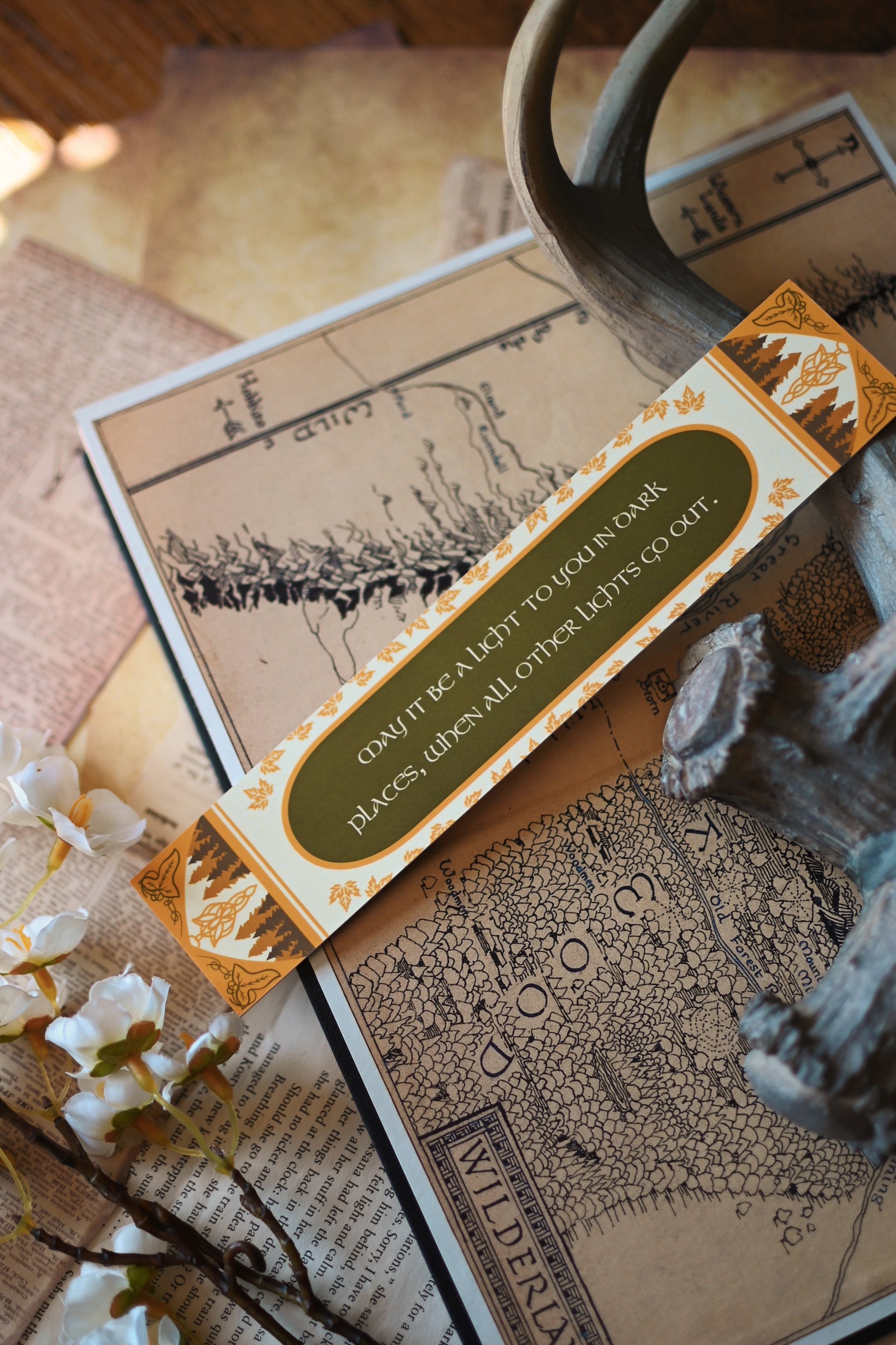 The Fellowship of the Ring inspired bookmark propped up against a background of the map of Middle Earth. The quote on the bookmark is: "May it be a light to you in dark places, when all other lights go out."