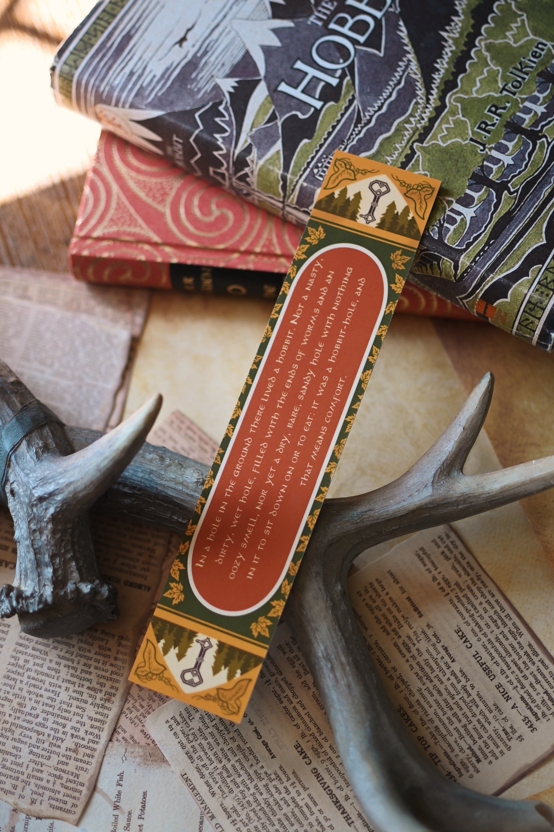 The Hobbit inspired bookmark featuring the beloved quote from the book with the bookmark resting against a copy of the book by J.R.R Tolkien. The quote is: "In a hole in the ground there lived a hobbit. Not a nasty, dirty, wet hole, filled with the ends of worms and an oozy smell, nor yet a dry, bare, sandy hole with nothing in it to sit down on or to eat: it was a hobbit-hole, and that means comfort."