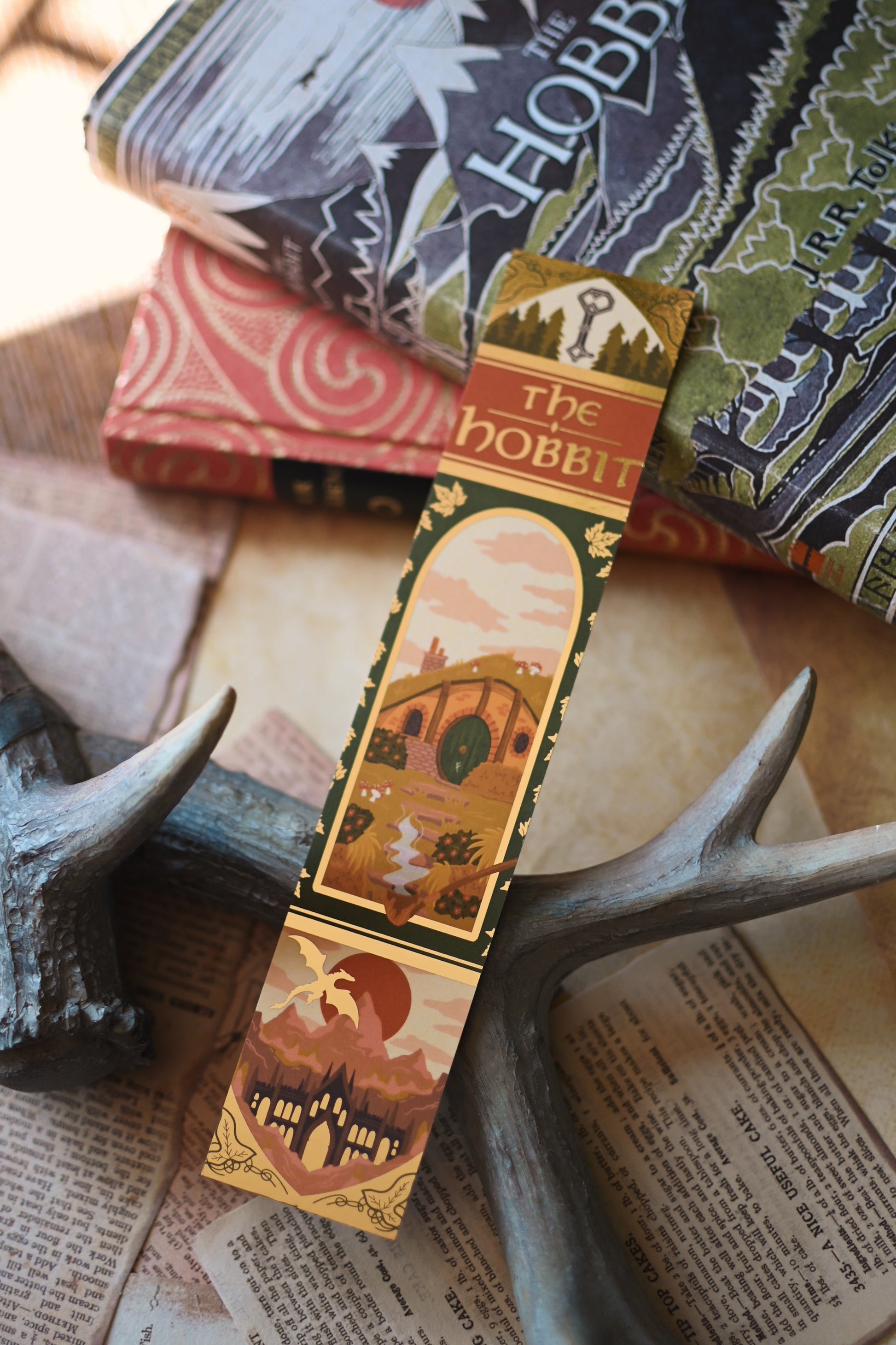 No One Designs foiled The Hobbit bookmark resting against copies of The Hobbit bby J.R.R Tolkien with the sunlight reflecting against the bookmark. In the background, there are book pages and the bookmark is being propped up against the book spines. 