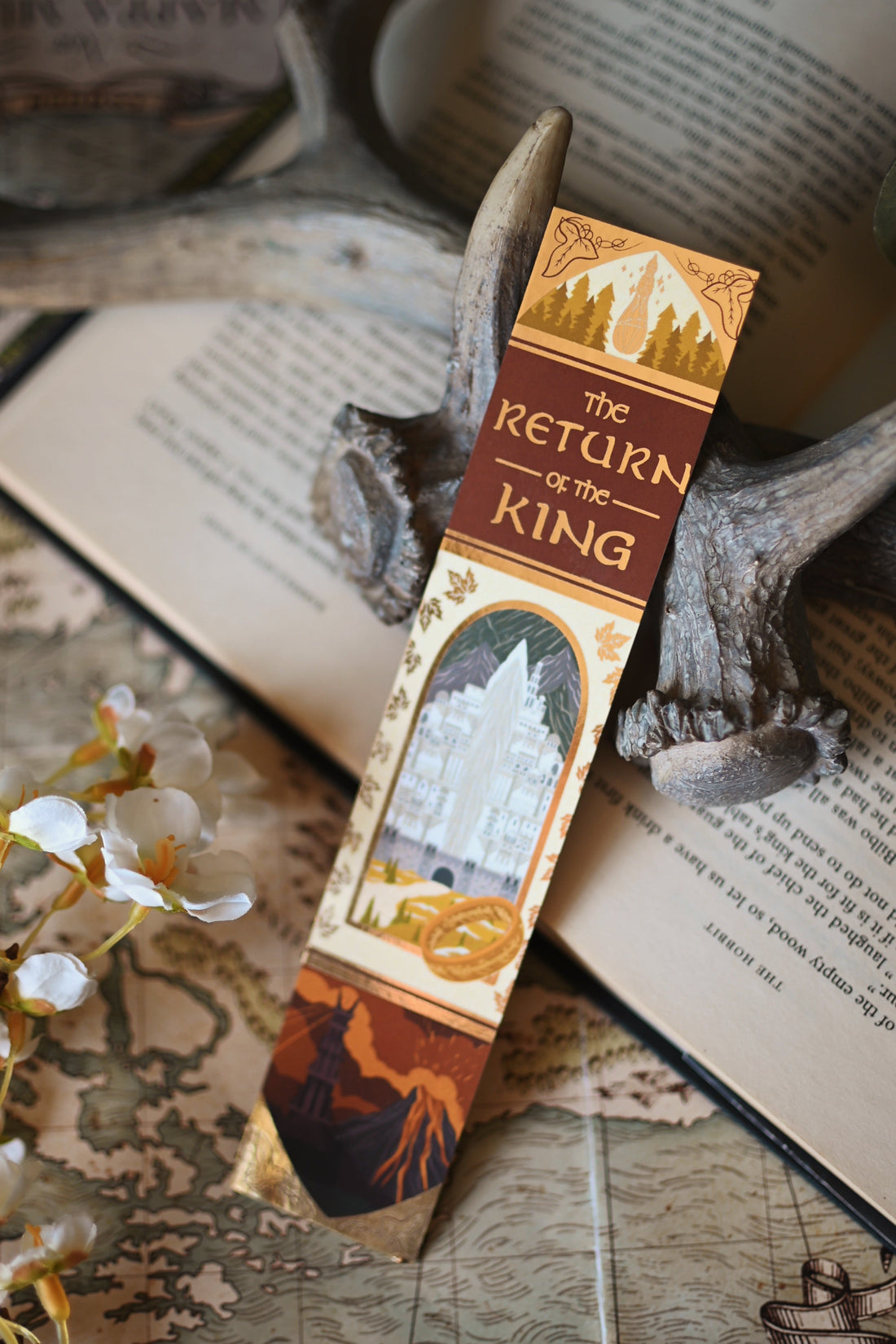 Propped up against a background of stained bookpages and orchid flowers, no one designs foiled bookmark inspired by J.R.R Tolkien's The Return of the King rests against an open copy of the book.