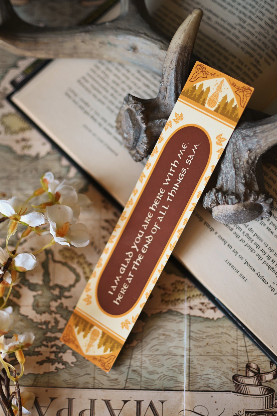 Propped up against a background of stained bookpages and orchid flowers, no one designs foiled bookmark inspired by J.R.R Tolkien's The Return of the King rests against an open copy of the book.