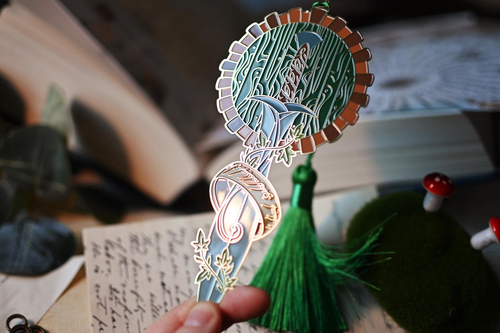 Chloe holding the tip of the Under the Hill inspired bookmark up to the light to show the intricate enamel detailing of the hobbit hole door, sting and the one ring. 